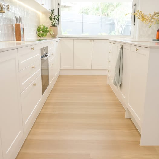 A bright, modern kitchen with white shaker-style cabinets, light wood flooring, and a large window in the background.