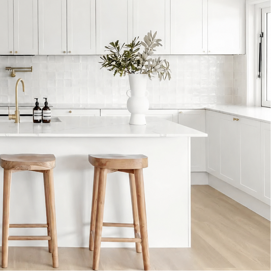 A bright, modern kitchen featuring white cabinets, a white island with a light marble countertop, three wooden bar stools, and a gold faucet. A white vase with greenery sits on the counter.
