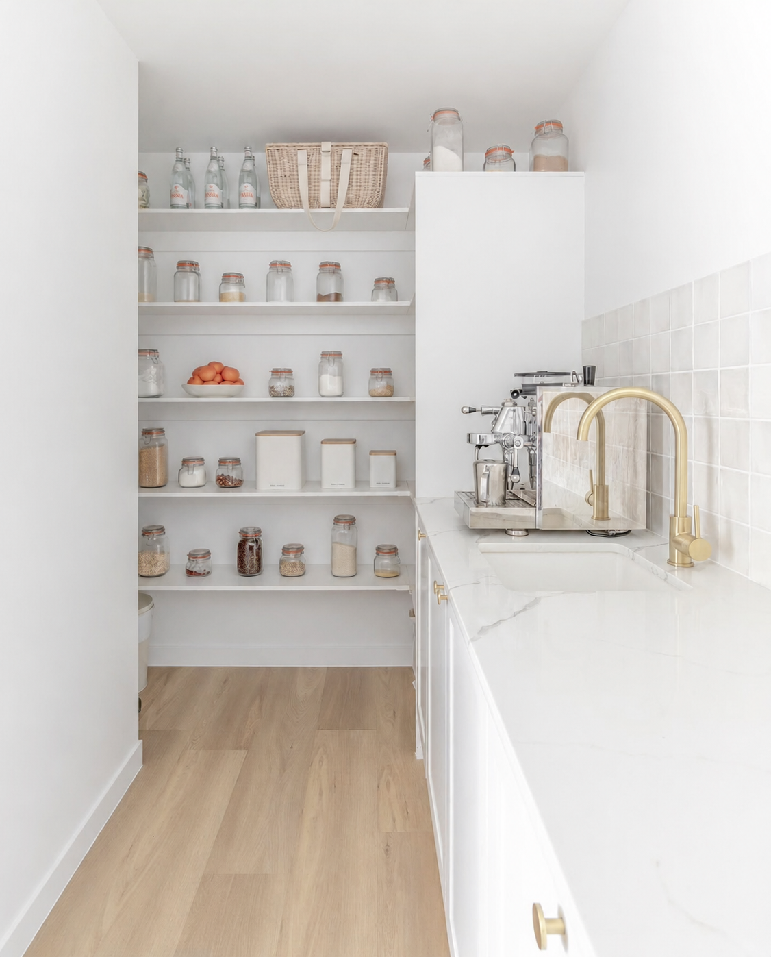 A bright, modern kitchen pantry with white shelves stocked with glass jars and canisters, a white marble countertop, a gold faucet, and a stainless steel espresso machine.