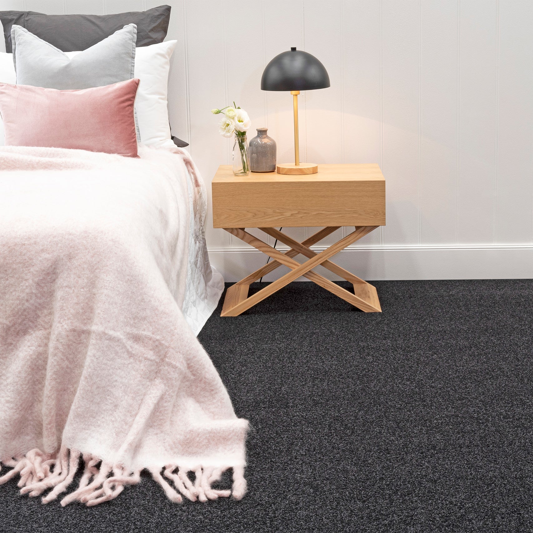 Bedroom with dark gray carpet, Pacific in Graphite. Bed with pink blanket and pillows, wooden nightstand with lamp.