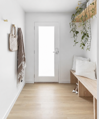 A bright, modern hallway with light wood flooring, a white door with a frosted glass panel, a wooden bench with white pillows on the right, and a woven tote bag and brown throw blanket hanging on the left wall.