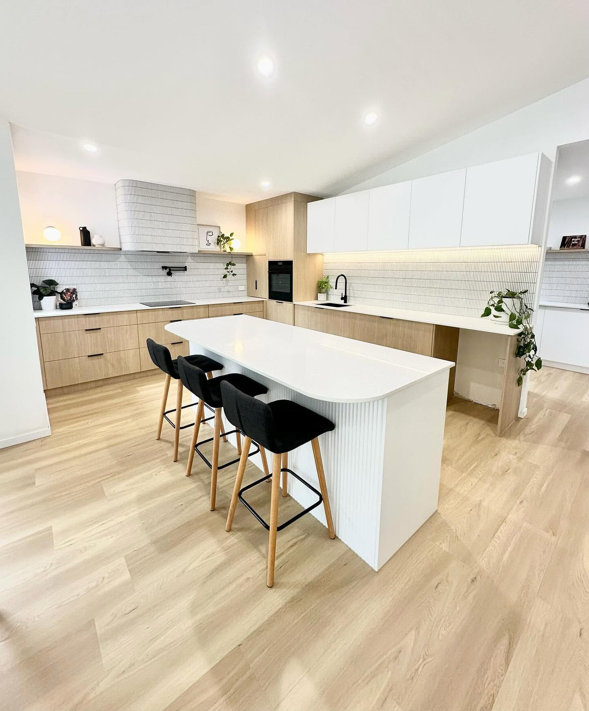 A modern kitchen island with a white ribbed base and curved white top, paired with three black bar stools featuring light wood legs, set in a bright kitchen with light wood flooring and cabinetry.