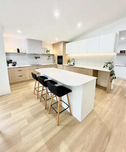 A modern kitchen island with a white ribbed base and curved white top, paired with three black bar stools featuring light wood legs, set in a bright kitchen with light wood flooring and cabinetry.