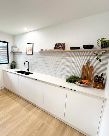 A modern kitchen corner with white cabinets, a white countertop, a black sink and faucet, a white subway tile backsplash, light wood floating shelves, and light wood flooring. Decorative items, plants, and fresh vegetables are arranged on the shelves and counter.