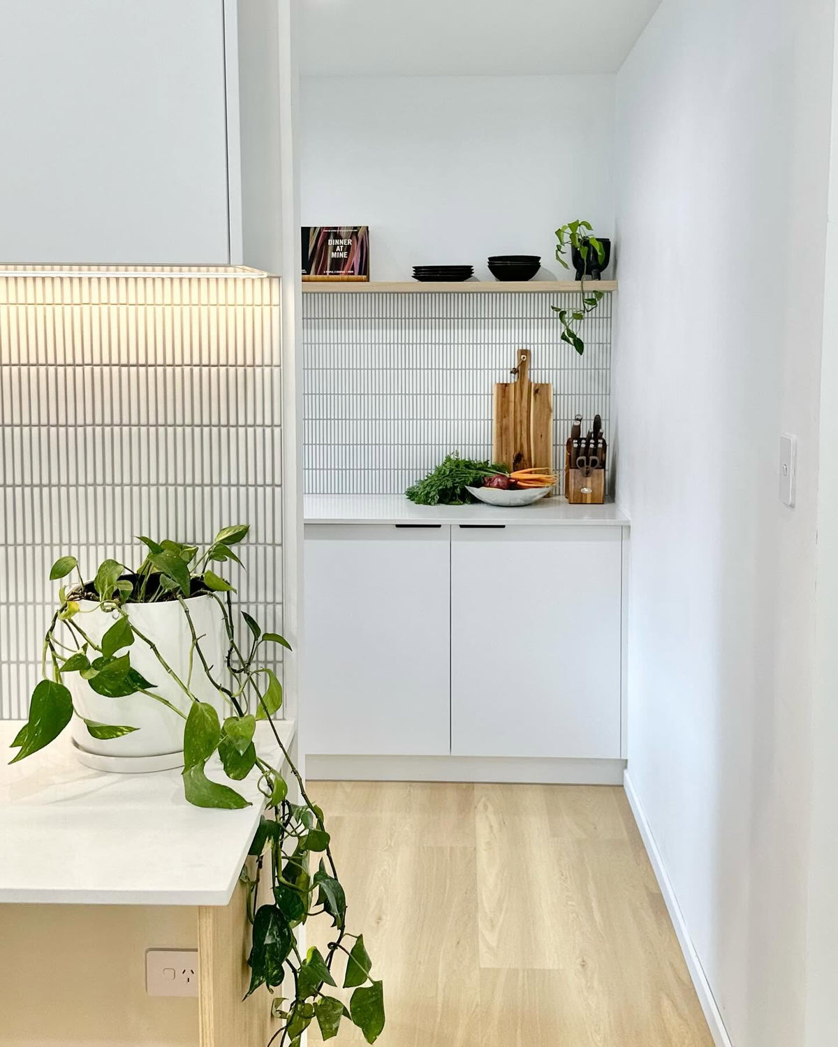 A modern kitchen corner with white cabinets, a white tiled backsplash, light wood flooring, open wooden shelves with small decor items, and a potted green plant in the foreground.