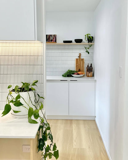 A modern kitchen corner with white cabinets, a white tiled backsplash, light wood flooring, open wooden shelves with small decor items, and a potted green plant in the foreground.